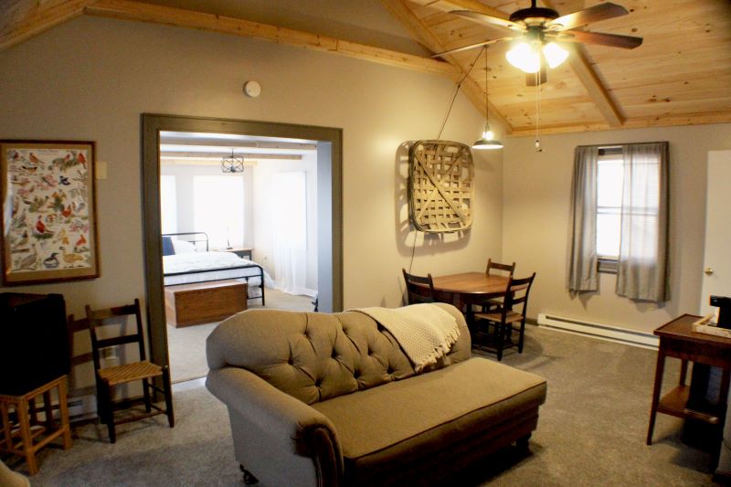 A rustic living space with a vaulted wooden ceiling, a beige tufted sofa, a round dining table, and a glimpse into an adjacent room.