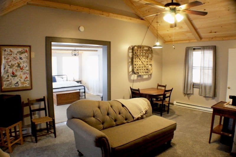 A rustic living space with a vaulted wooden ceiling, a beige tufted sofa, a round dining table, and a glimpse into an adjacent room.