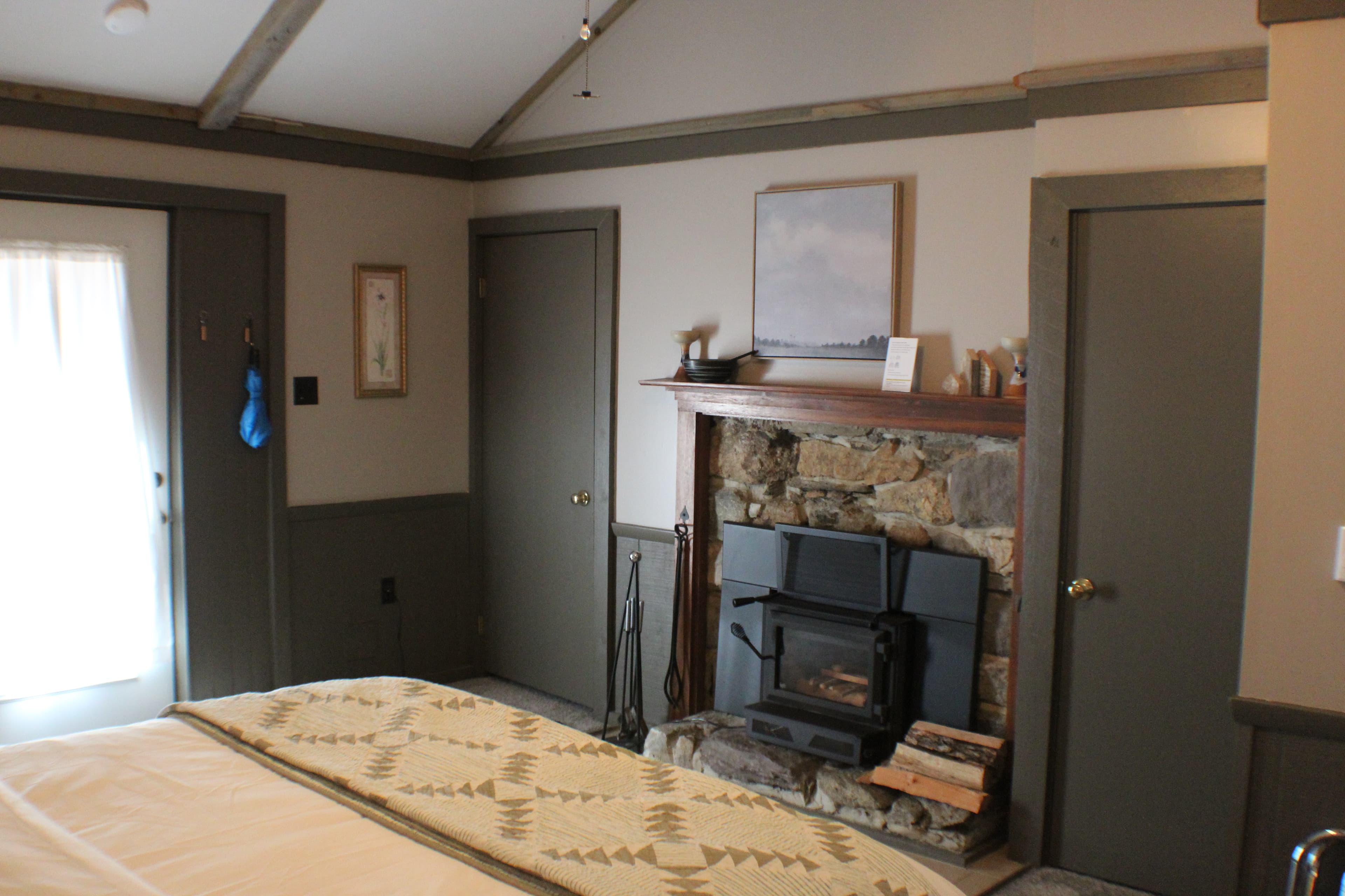 A rustic bedroom featuring a stone fireplace with a wood-burning stove, a bed with a patterned throw, and a door leading outside.