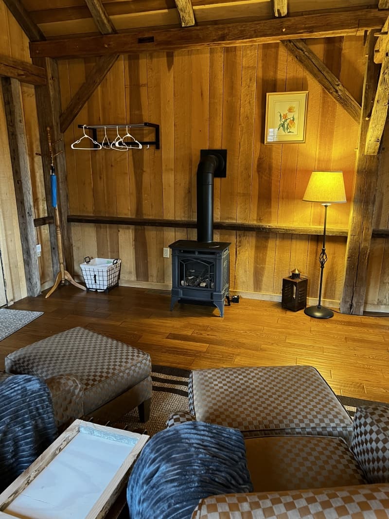 A cozy corner of a room with a wood-paneled wall, exposed beams, a black wood-burning stove, and upholstered seating in the foreground.