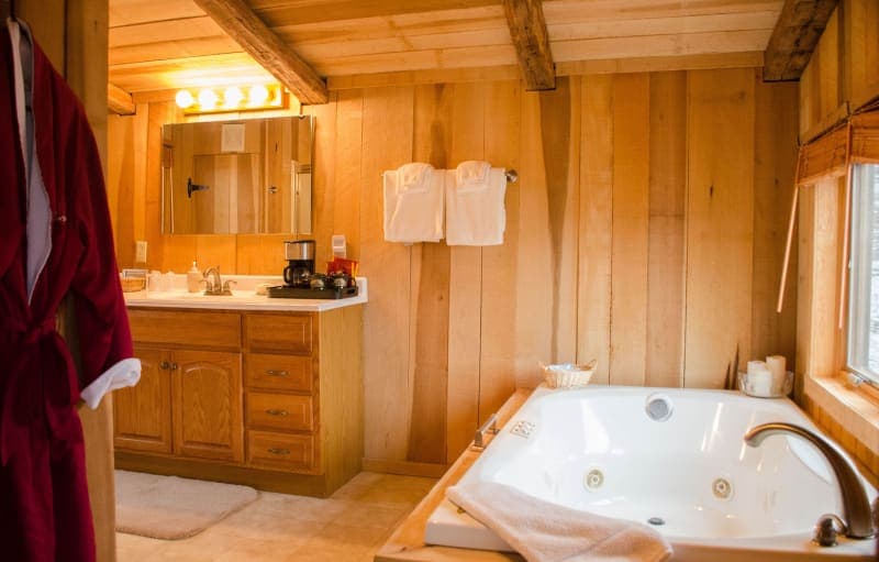 A rustic bathroom featuring warm wood paneling, a large jetted tub, a vanity with a double sink, and a red bathrobe hanging nearby.