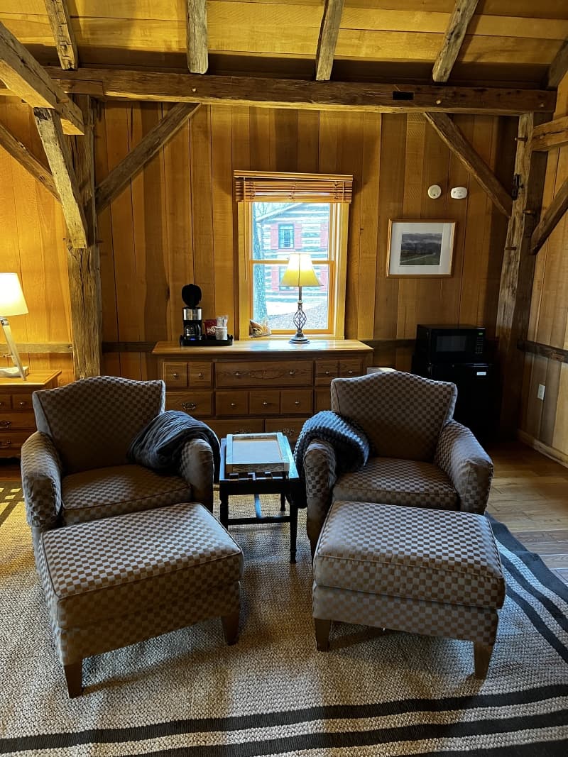 A comfortable seating area in a wood-paneled room with exposed beams, featuring two armchairs with ottomans, a small table, and a dresser with a lamp.