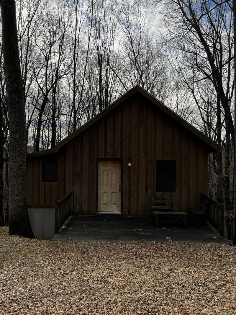 A rustic brown cabin with a white door and a small wooden bench, nestled amongst bare trees on a gravel path.