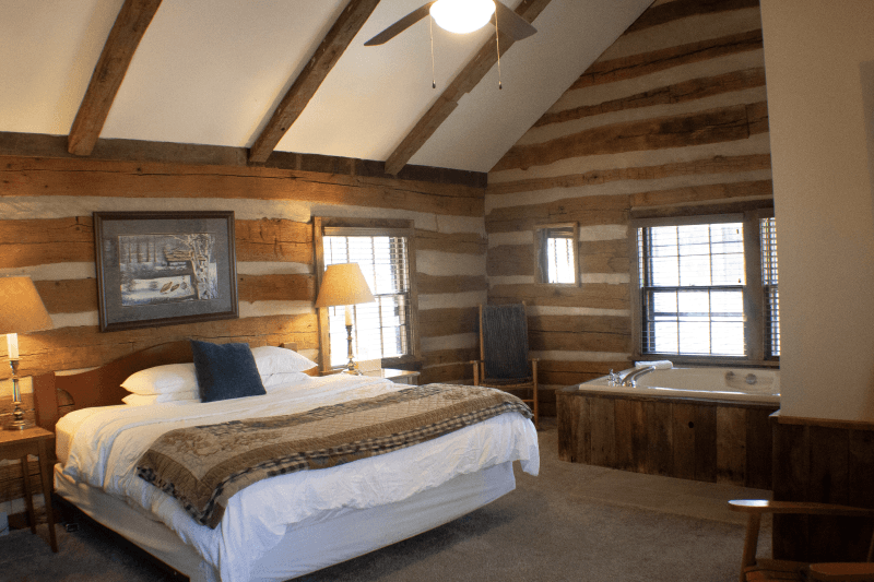 A rustic log cabin bedroom featuring a king-sized bed, a jetted tub, exposed log walls, and a vaulted ceiling with a fan.