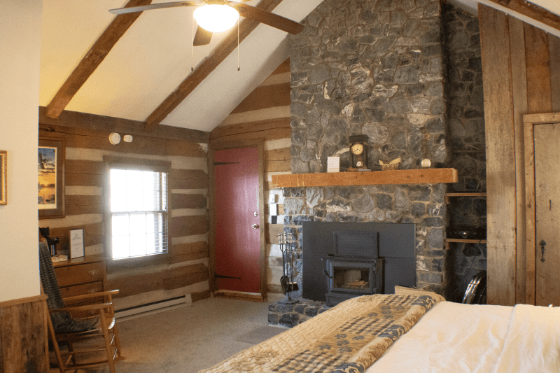 A rustic log cabin interior with a stone fireplace, a wood-burning stove, a red door, and a vaulted ceiling with a fan.
