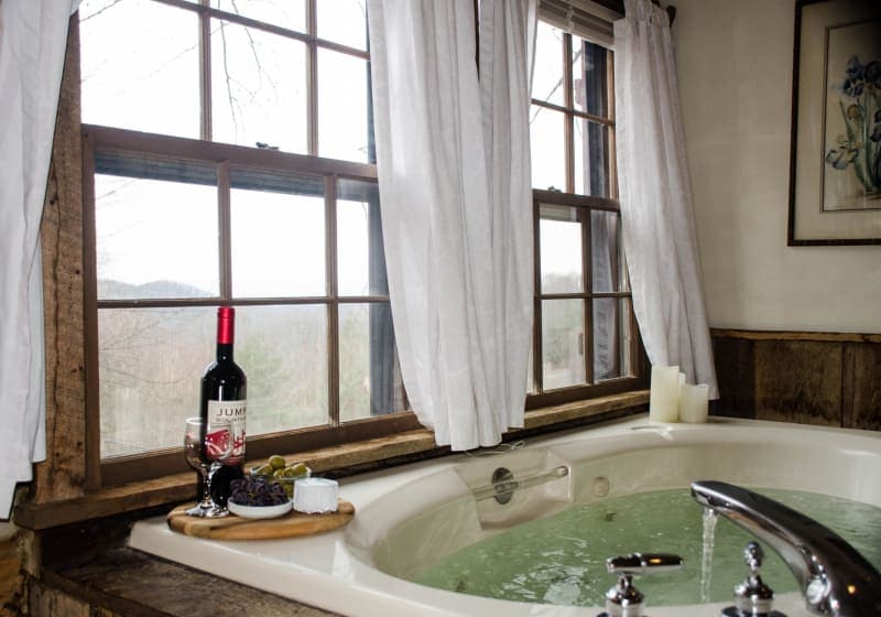 A luxurious jetted bathtub by a window with a scenic view, accompanied by a tray holding a bottle of wine, a glass, and snacks.
