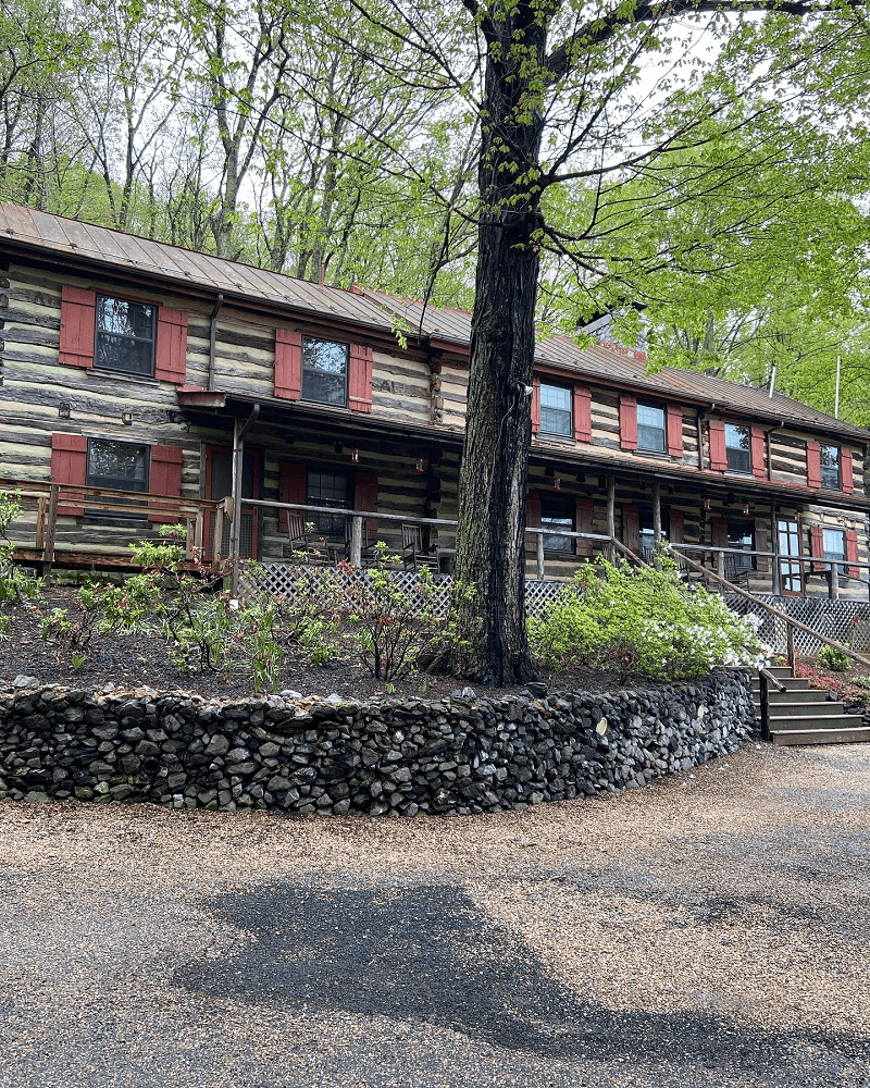 A two-story rustic lodge with red window shutters and a porch, nestled among trees, with a stone wall in the foreground.
