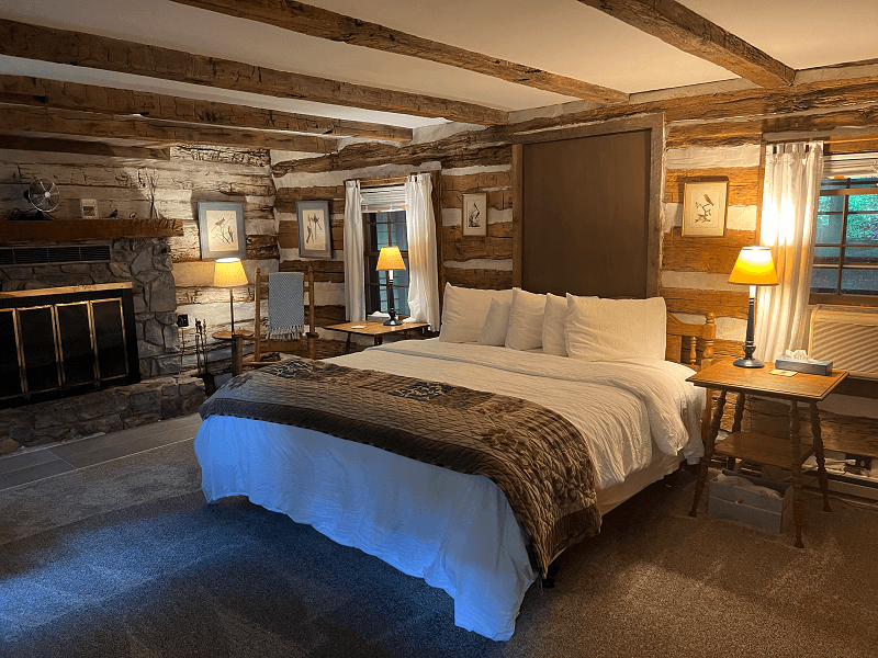 A warm and inviting bedroom in a log cabin, featuring a king bed, a stone fireplace, exposed wooden beams, and soft lamp lighting.