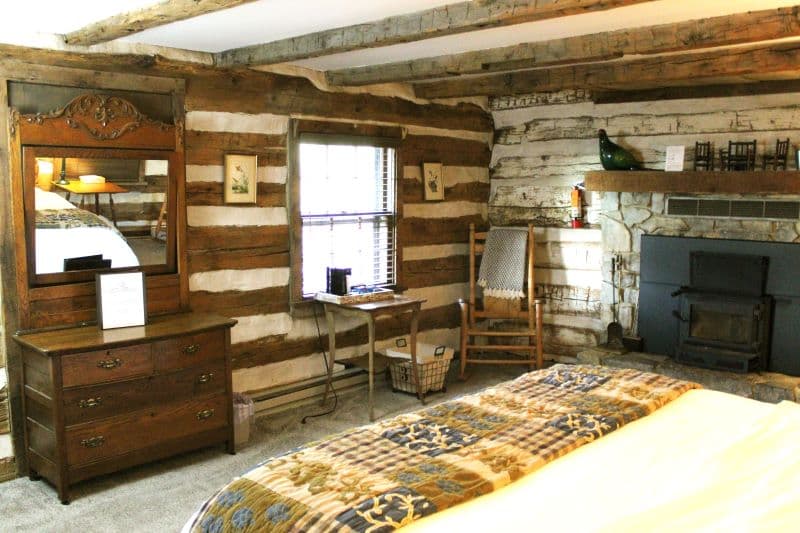 A rustic log cabin bedroom with a stone fireplace, a wood-burning stove, a wooden dresser with a mirror, and a rocking chair.