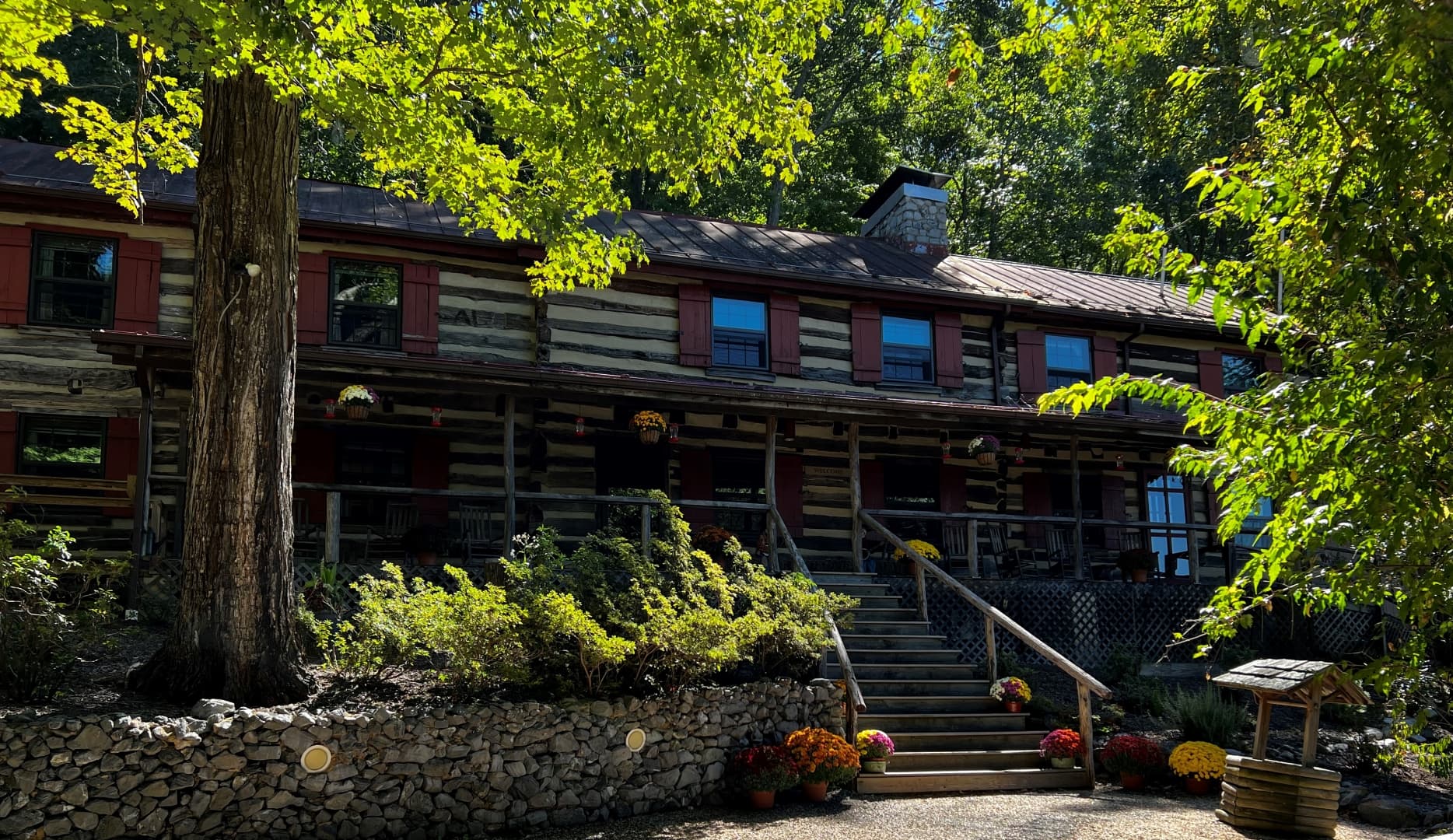 A multi-story log cabin lodge with a wide front porch and red window frames, surrounded by lush green trees and a stone retaining wall.