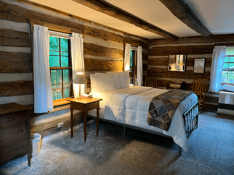 A rustic log cabin bedroom with a queen bed, white bedding, a colorful throw, two nightstands with lamps, and multiple windows.