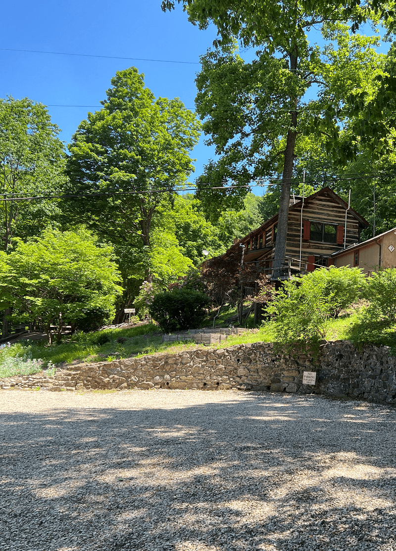 A gravel parking area leads to a rustic log cabin nestled among lush green trees, with a stone retaining wall and various plants.