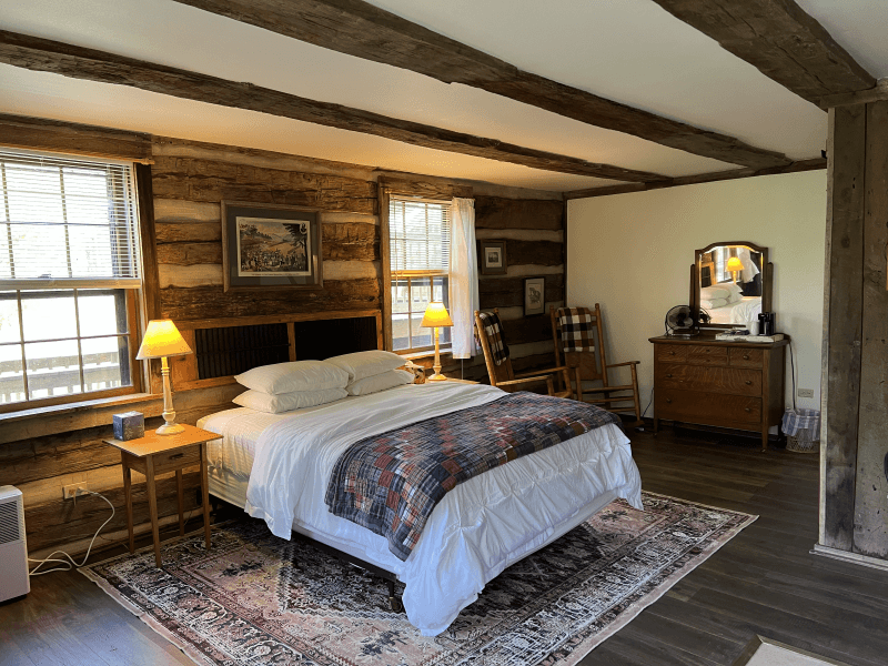 Queen size bed set in an old log building with exposed logs on walls and ceiling and high end luxury vinyl plank pet friendly flooring.