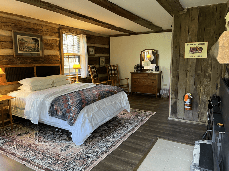 Another view of the queen size bed set against 200 year old log cabin walls and a wood burning fireplace with stone surround.
