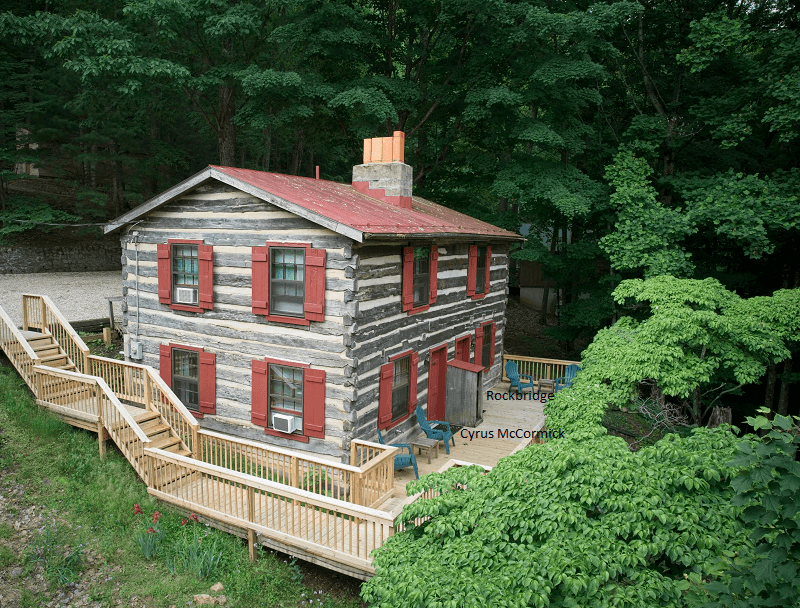 A quaint two-story log cabin with a red roof and red shutters, featuring a wraparound deck and a long wooden staircase leading down.