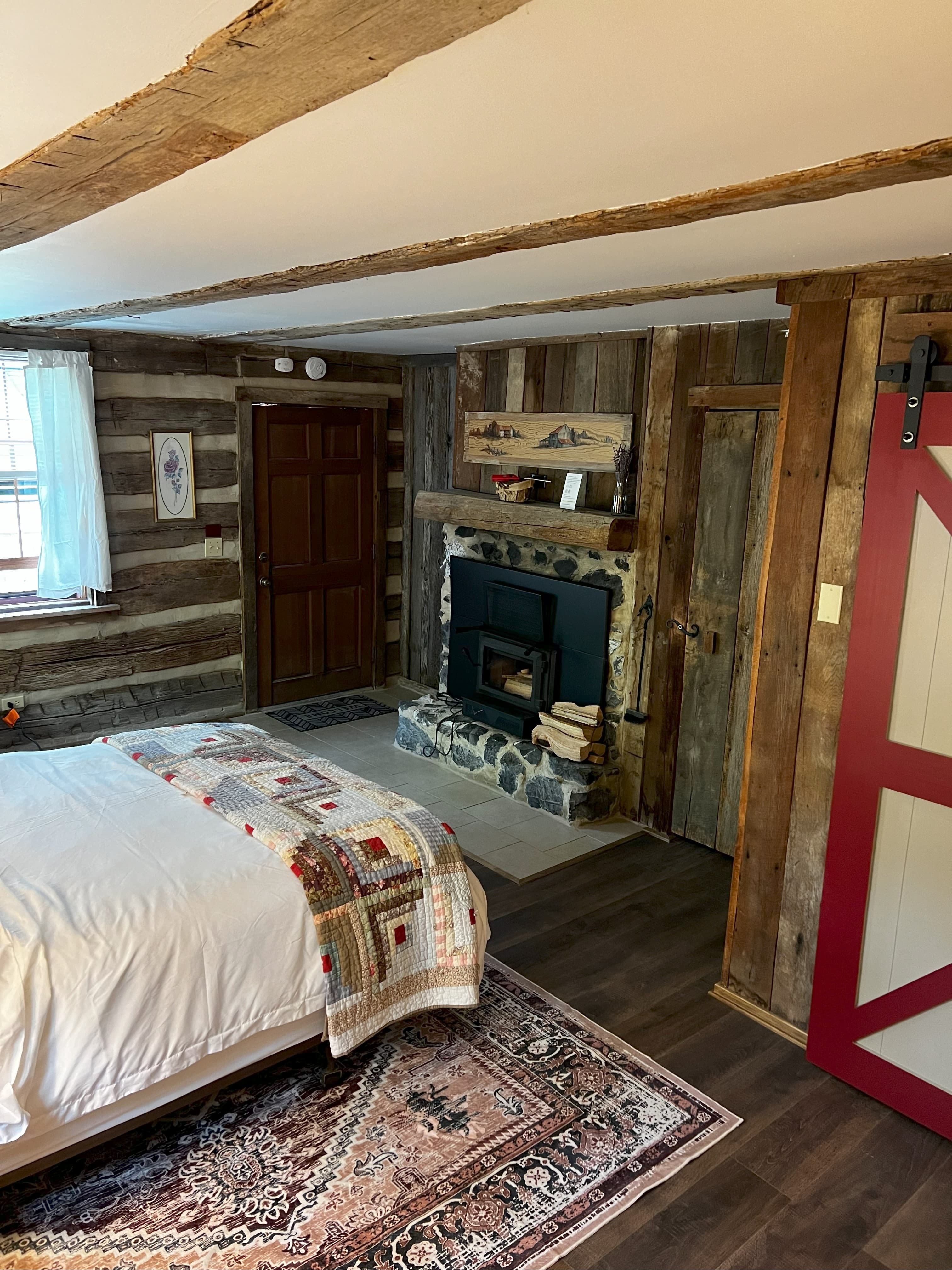 A cozy log cabin bedroom featuring a queen bed, a stone fireplace with a wood-burning stove, exposed beams, and a red barn-style sliding door.