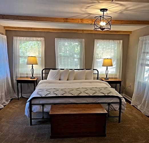 A bedroom with a king-sized bed, white linens, a patterned throw, two nightstands with lamps, and several windows with sheer curtains.
