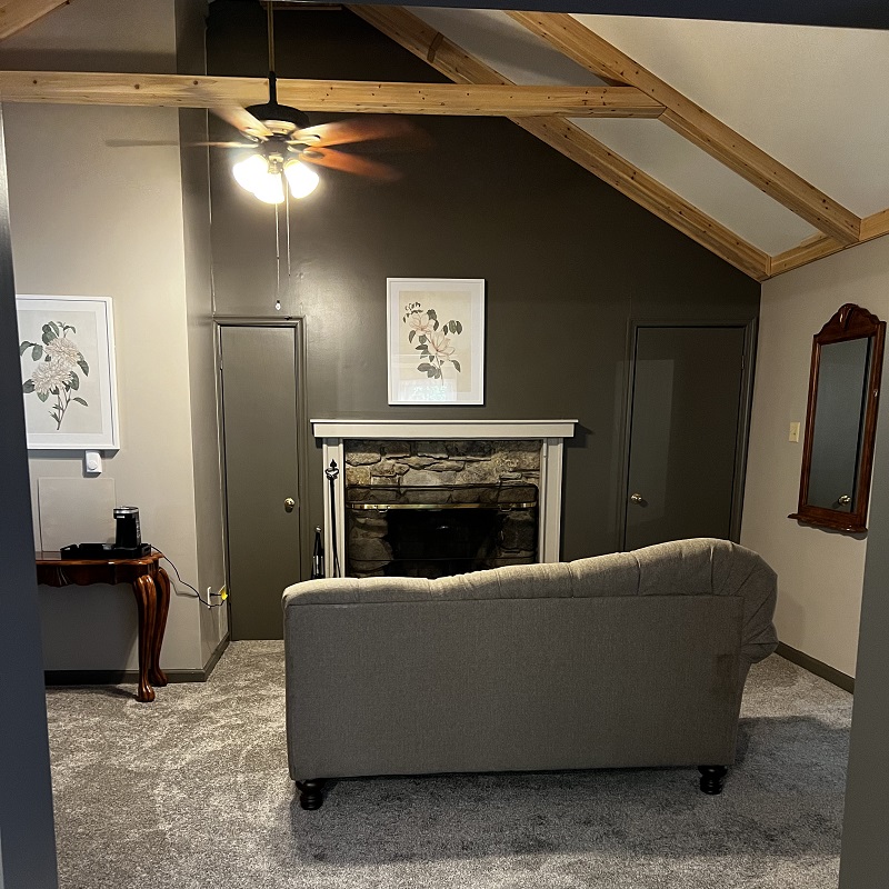 A rustic living room with a vaulted ceiling, exposed beams, a stone fireplace, a ceiling fan, and a gray sofa.