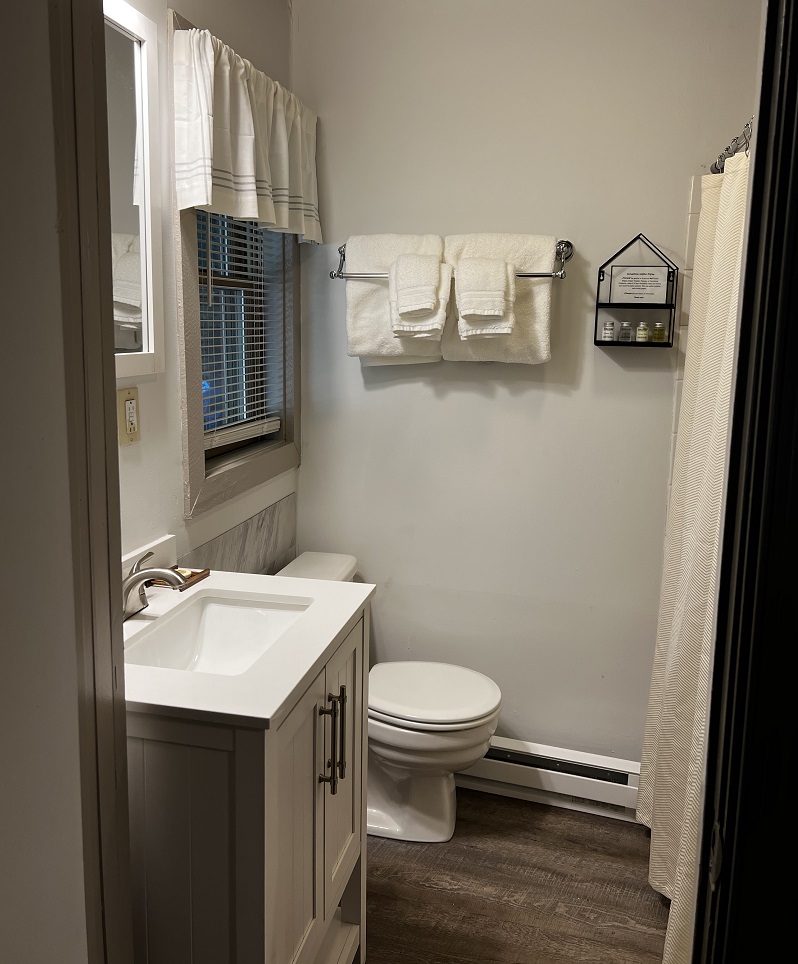 A small, well-lit bathroom with a white vanity, a toilet, a shower with a white curtain, and white towels hanging on a rack.