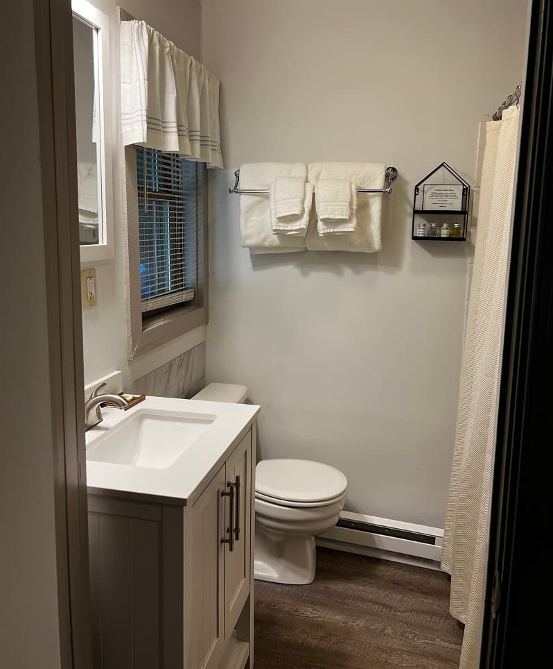 A small, well-lit bathroom with a white vanity, a toilet, a shower with a white curtain, and white towels hanging on a rack.