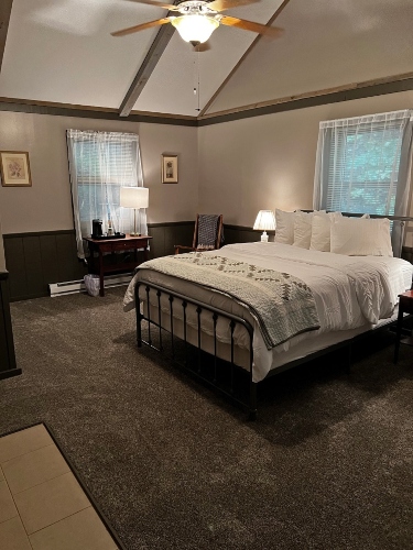 A cozy bedroom featuring a queen bed with white bedding, a gray throw, two nightstands with lamps, and a ceiling fan on a vaulted ceiling.