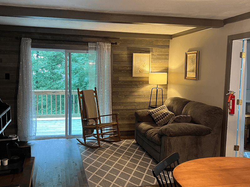A cozy living room featuring a sliding glass door to a deck, a wooden rocking chair, a dark sofa, and a patterned rug.