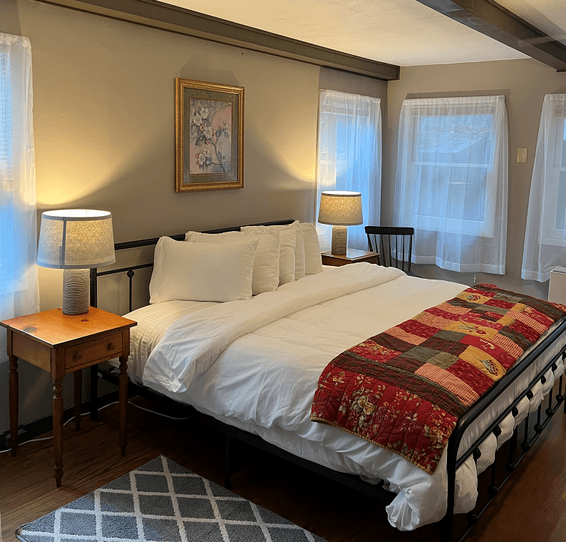 A well-lit bedroom with a king-size bed, white bedding, a colorful throw, two nightstands with lamps, and multiple windows.