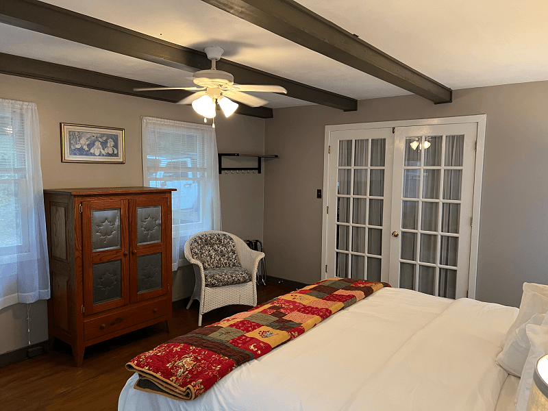 A bright bedroom featuring a king bed with a colorful throw, a wooden armoire, a wicker chair, and French doors.