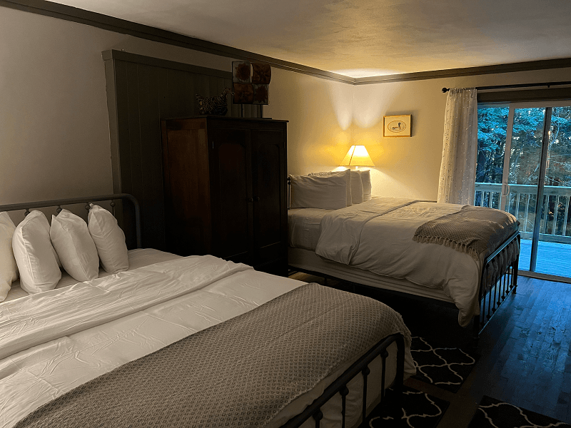 A spacious bedroom with two queen beds, each with white linens and a gray throw, a dark armoire, and a sliding door leading to a balcony.