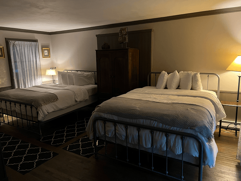 A softly lit bedroom featuring two beds with white and gray bedding, a dark wooden armoire, and two lamps.