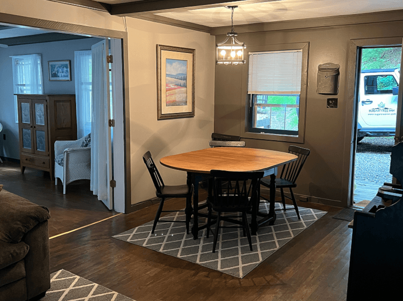 A dining area featuring a wooden drop-leaf table with four chairs, a geometric rug, a window, and a door leading outside.
