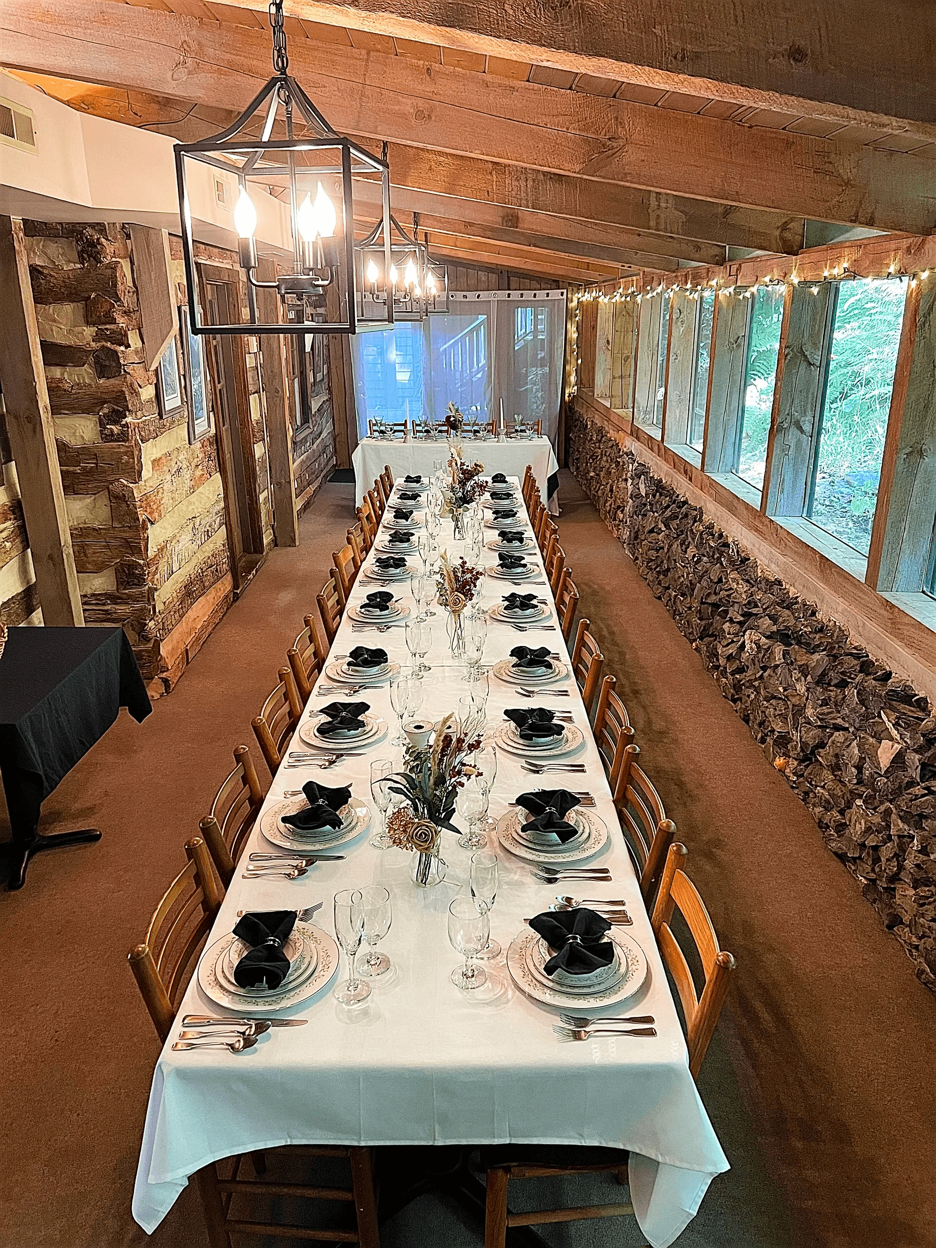 A long dining table is elegantly set with black napkins and tableware beneath wooden beams and soft lighting.