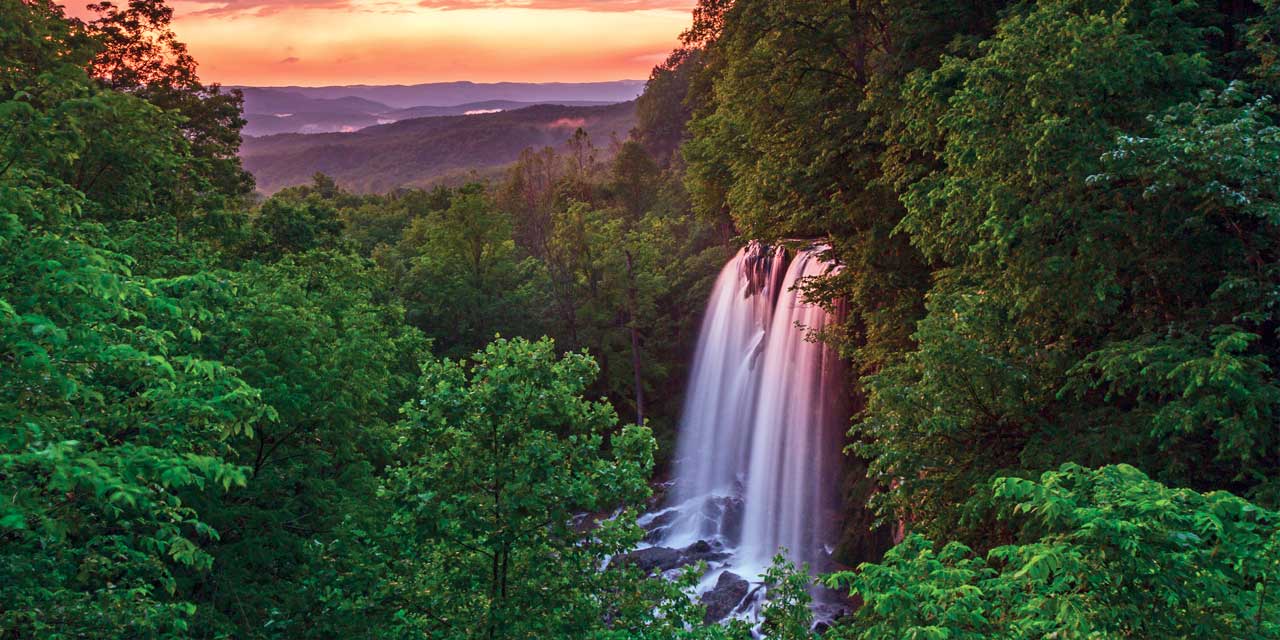 A tranquil waterfall cascades through lush green trees at sunset.