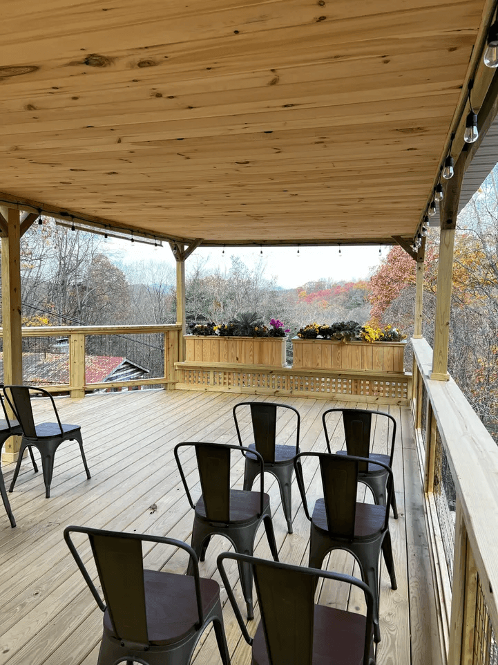 A spacious wooden deck with black metal chairs and a flower planter overlooking a treed landscape.