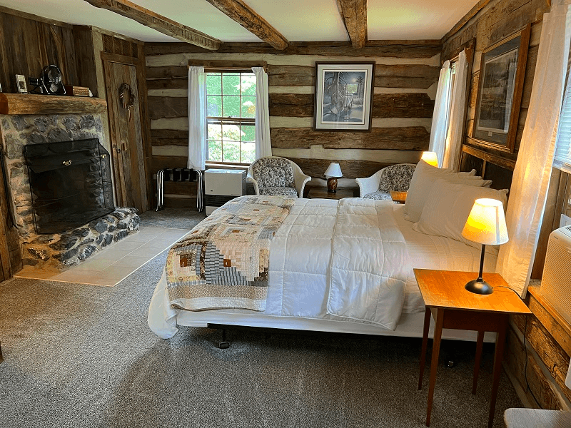 A cozy log cabin bedroom featuring a king-sized bed, two chairs, a stone fireplace, and large windows.