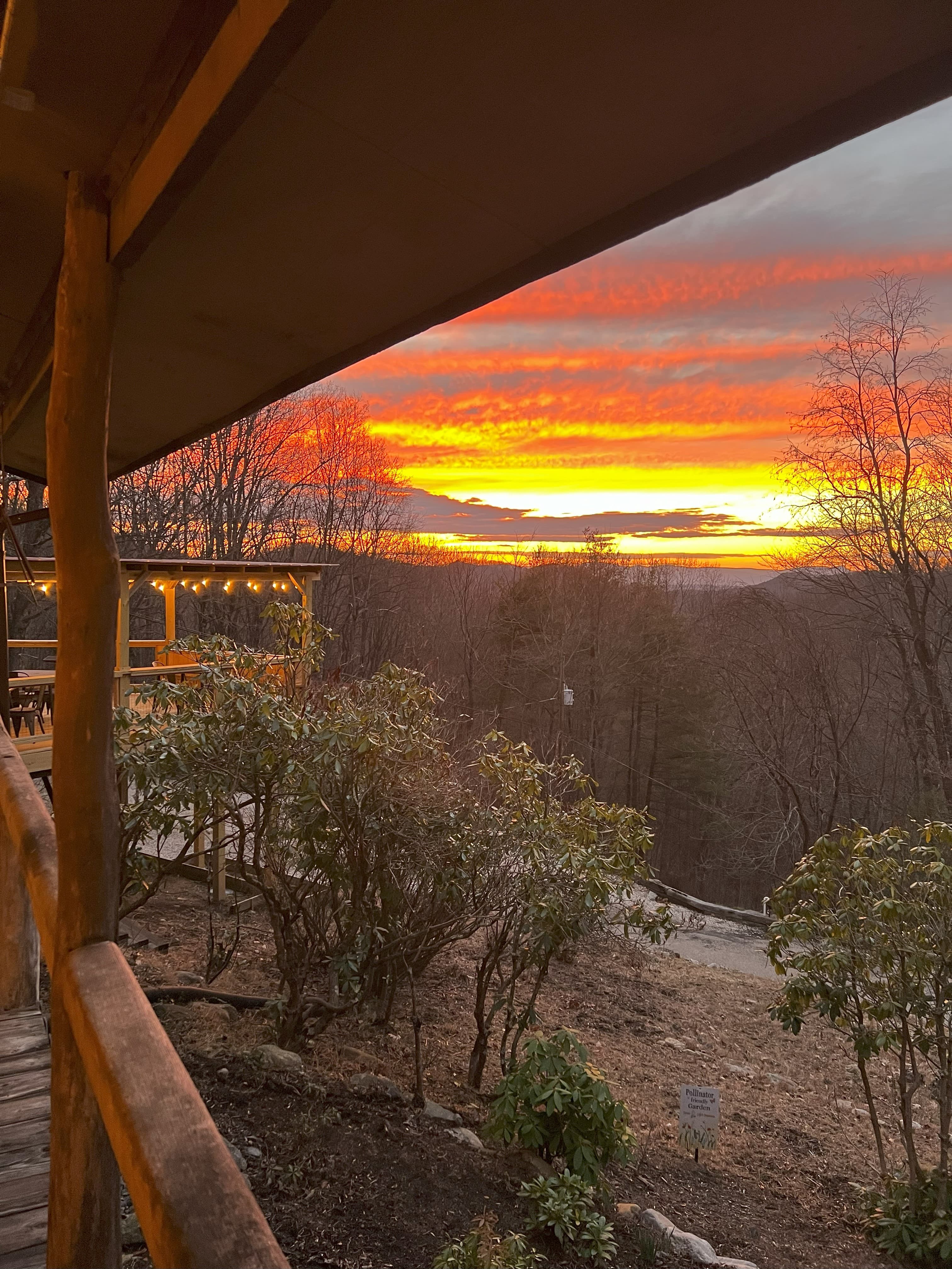 A vibrant sunset illuminates the sky over a wooded landscape, viewed from a cozy cabin porch.