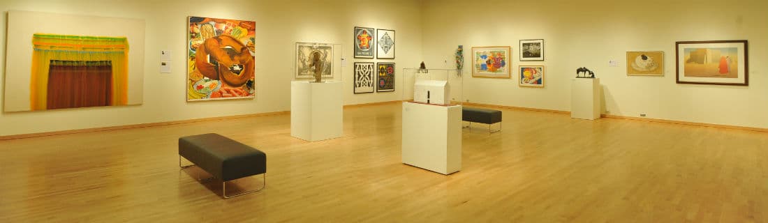 A spacious art gallery featuring various colorful paintings and sculptures displayed on white pedestals and walls.