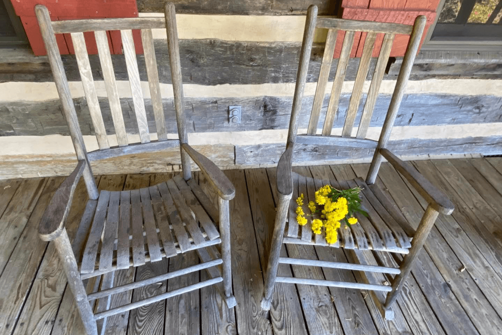 Two rustic rocking chairs sit on a wooden porch, adorned with a small bouquet of yellow flowers.