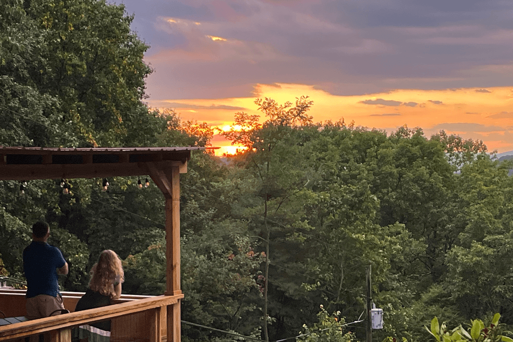 Two people on a porch looking out as the sun sets behind tall trees