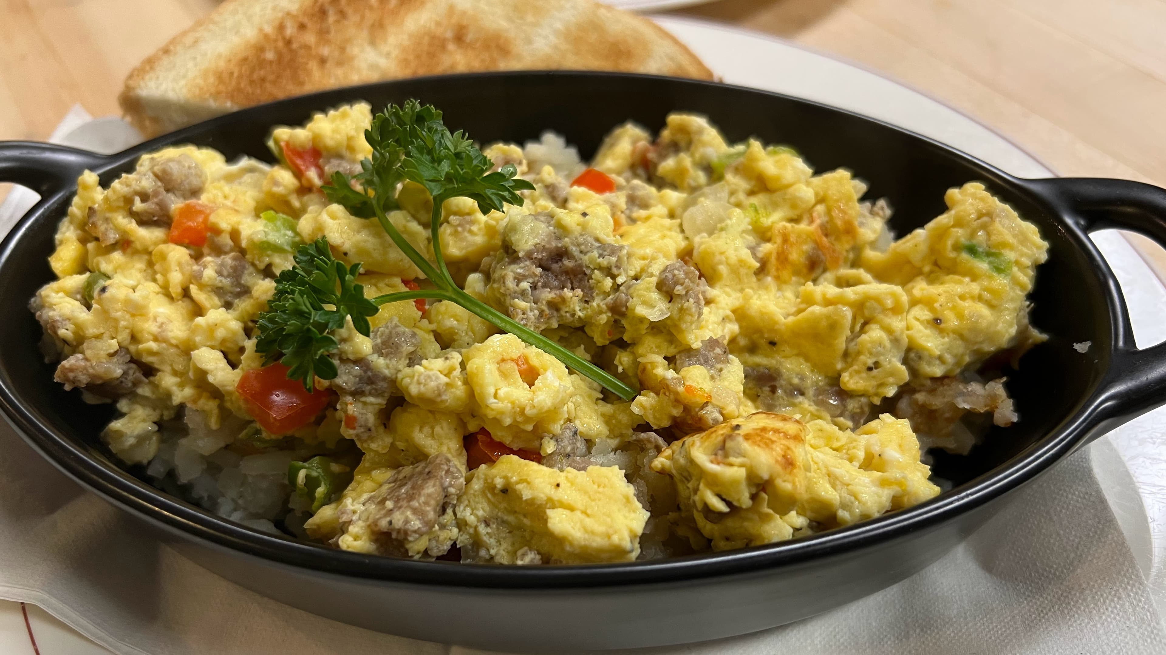 A skillet of scrambled eggs with sausage, vegetables, and a slice of toast.