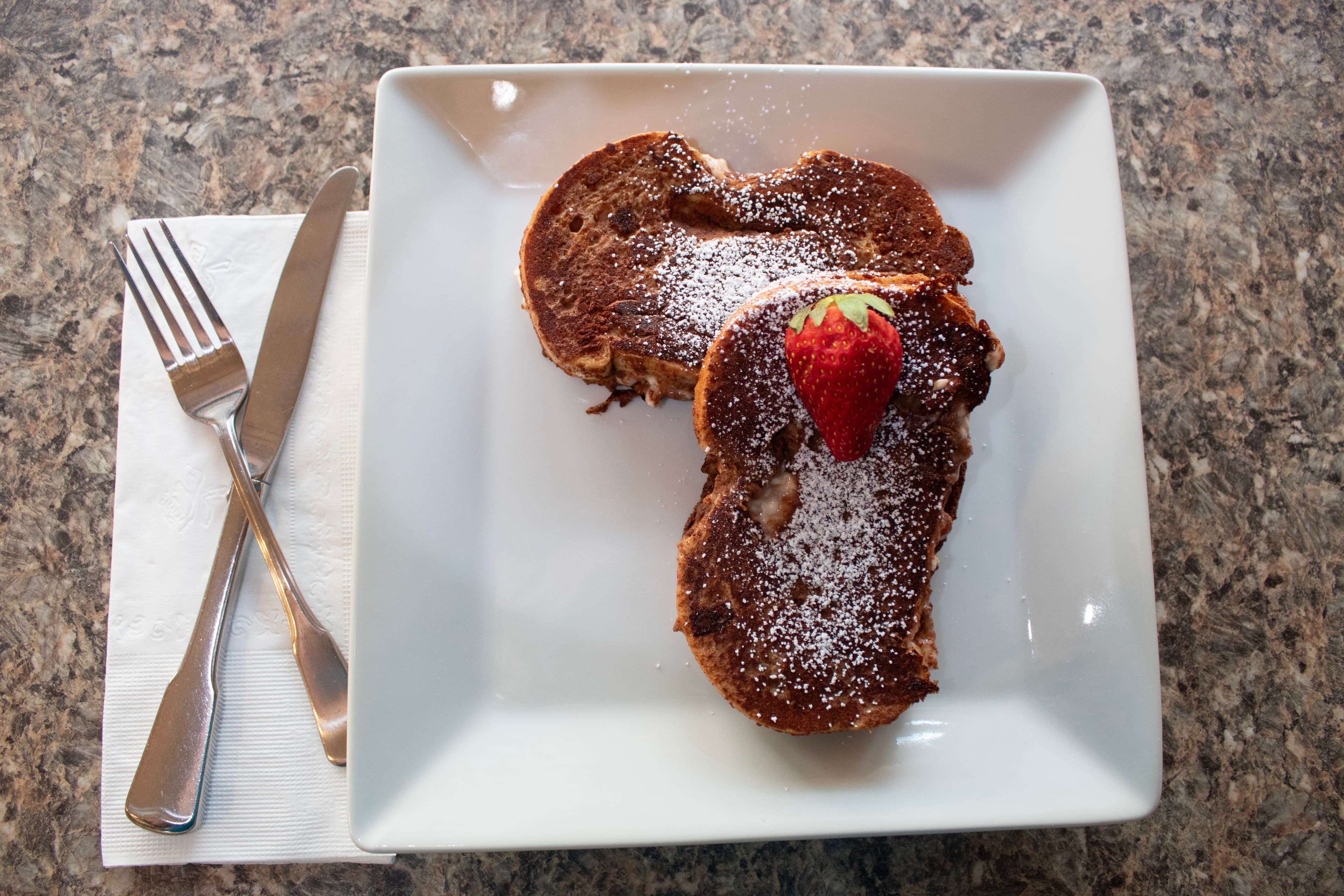 Two pieces of French toast topped with powdered sugar and a strawberry on a white plate, next to a knife and fork.