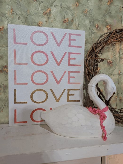 A decorative print with the word "LOVE" in various colors next to a white ceramic swan adorned with a pink ribbon, set against a floral wallpaper background.