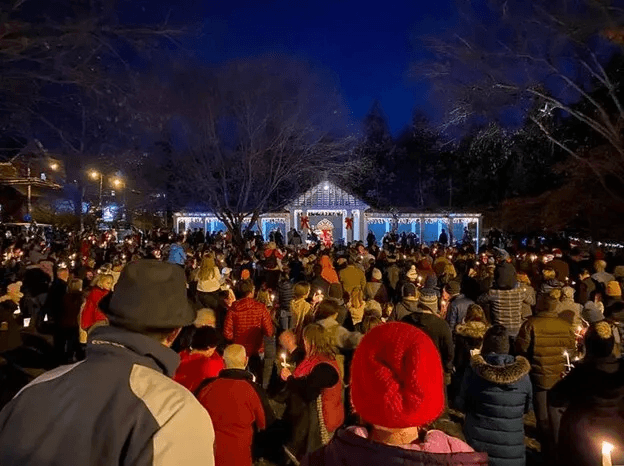 A large crowd gathers in a park at night, holding candles in front of a festive house adorned with lights.