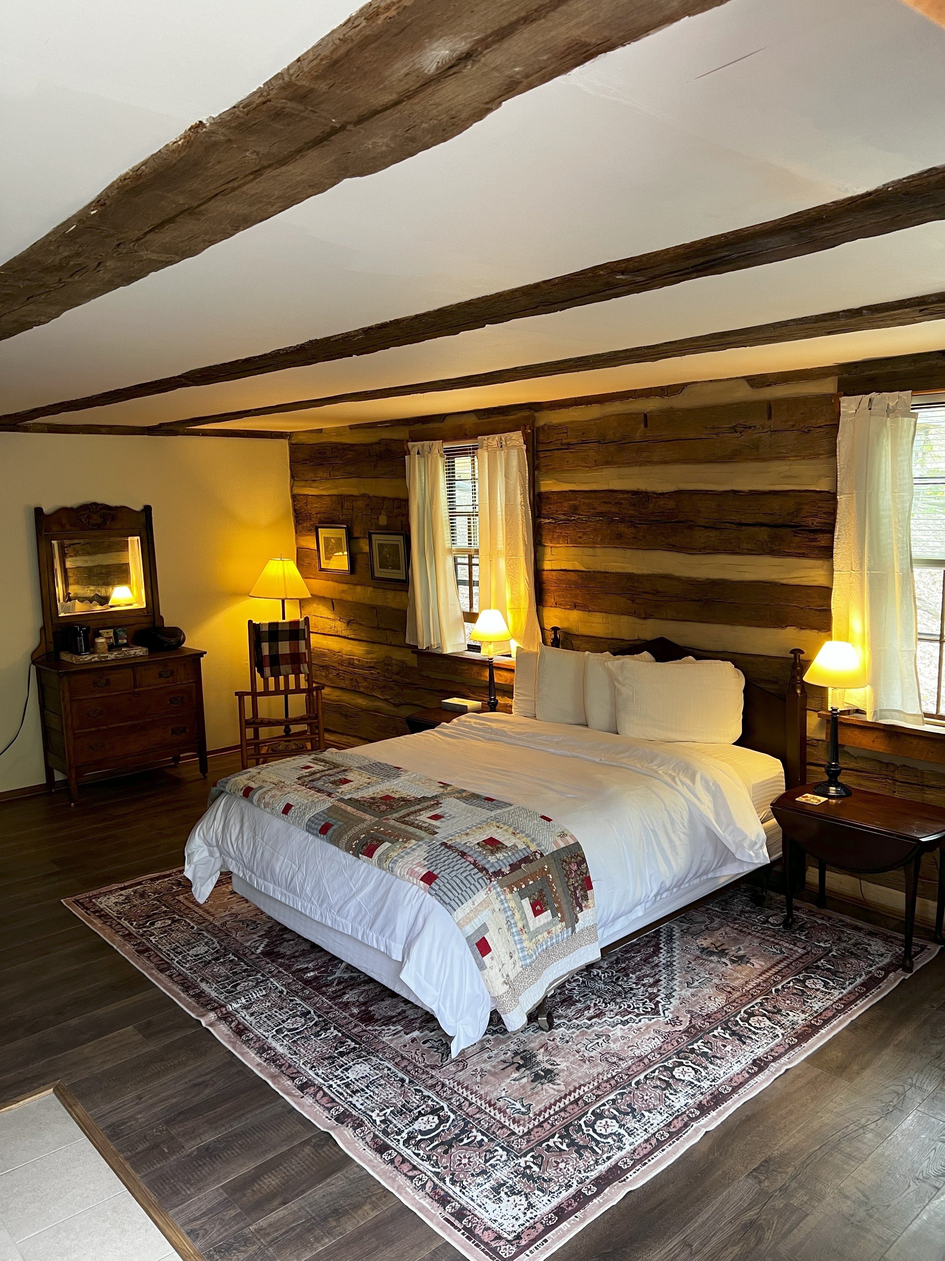 A charming log cabin bedroom with a queen bed, white bedding, a patterned throw, wooden walls, exposed beams, and two bedside lamps.