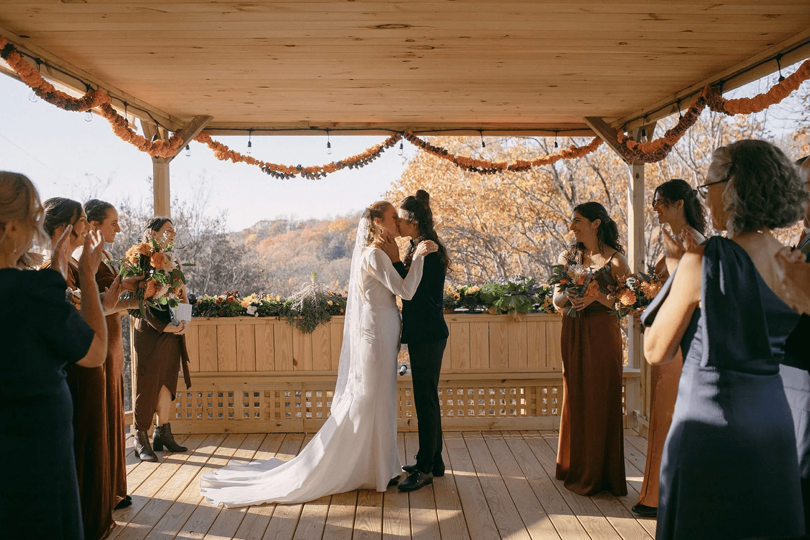 Two brides share a kiss during their outdoor wedding ceremony, surrounded by smiling guests.