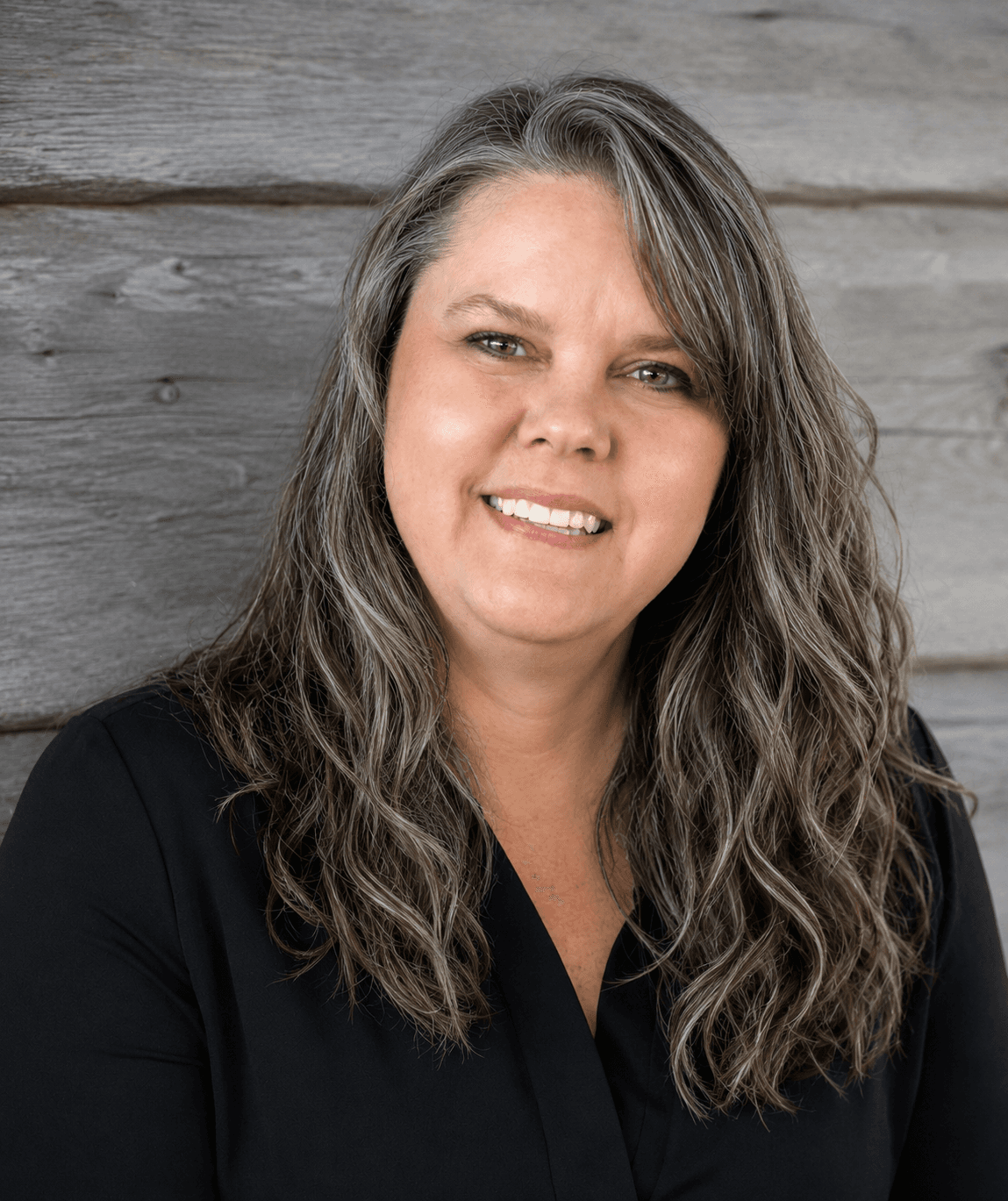 A woman with long, wavy gray hair smiles in front of a wooden background.