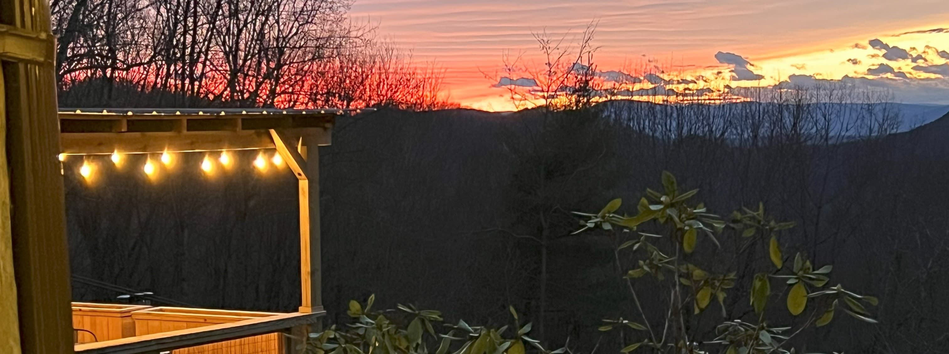 A colorful sunset over mountains, viewed from a wooden deck illuminated by string lights.