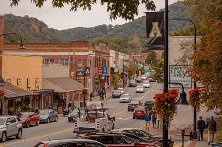 A bustling street in a small town featuring shops, parked cars, and surrounding hills.