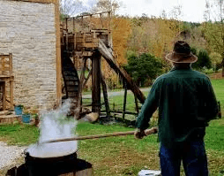 Man in green shirt over steaming pot of apple butter in front of stone flour mill