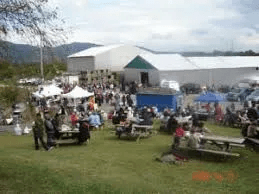 picnic tables set up in field with white barn in background and white tents with people walking around